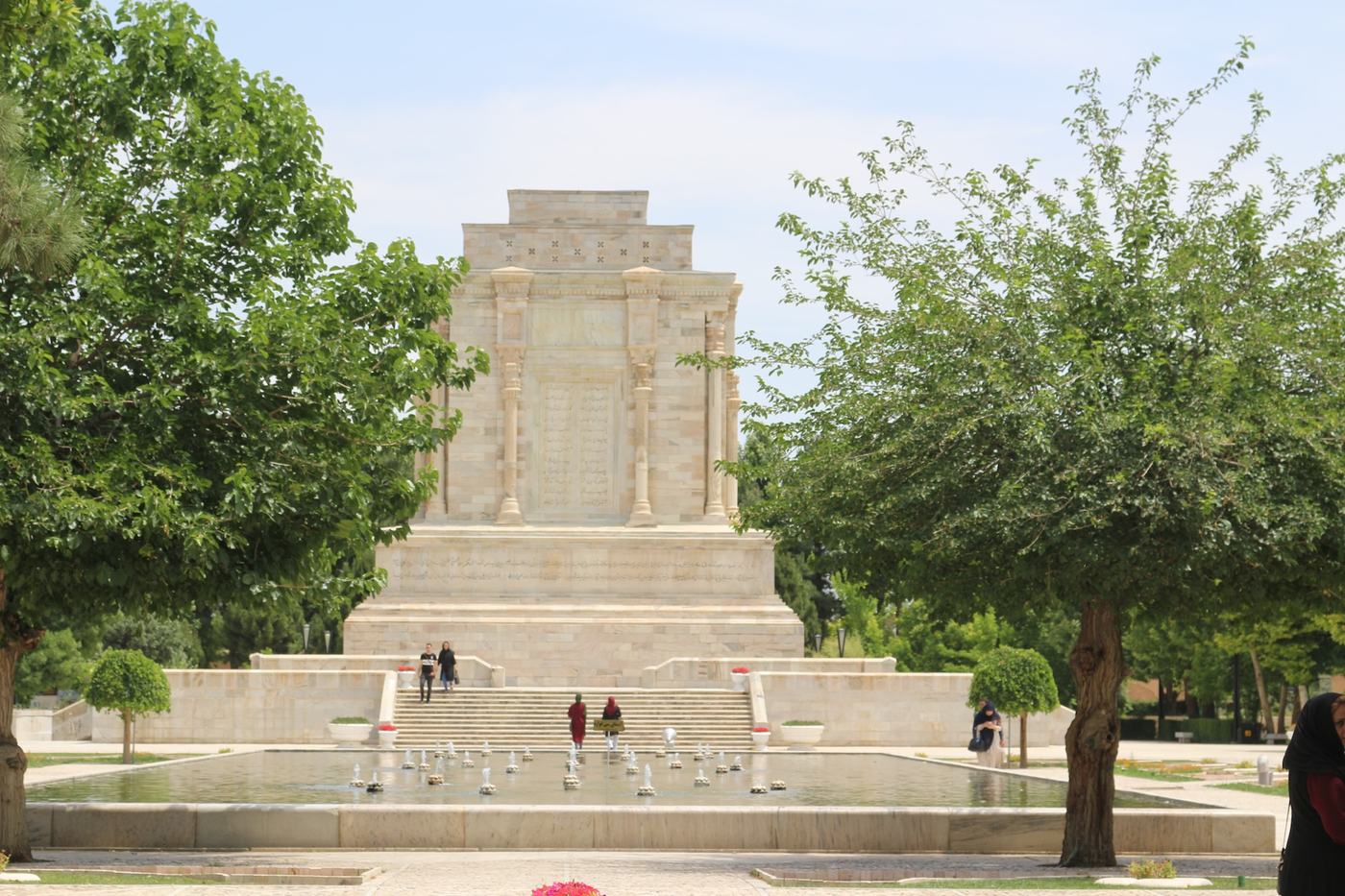 Front view of Ferdowsi’s mausoleum in Tus, a white stone monument surrounded by trees, fountains, and visitors walking through the garden.