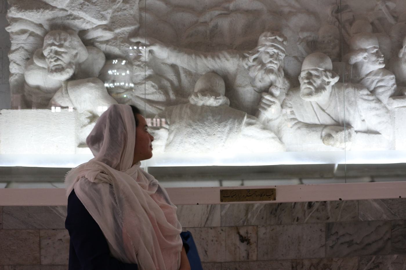 Author wearing a light headscarf inside Ferdowsi’s mausoleum in Tus, looking at a large stone relief depicting a scene from the Shâh Nâmeh.